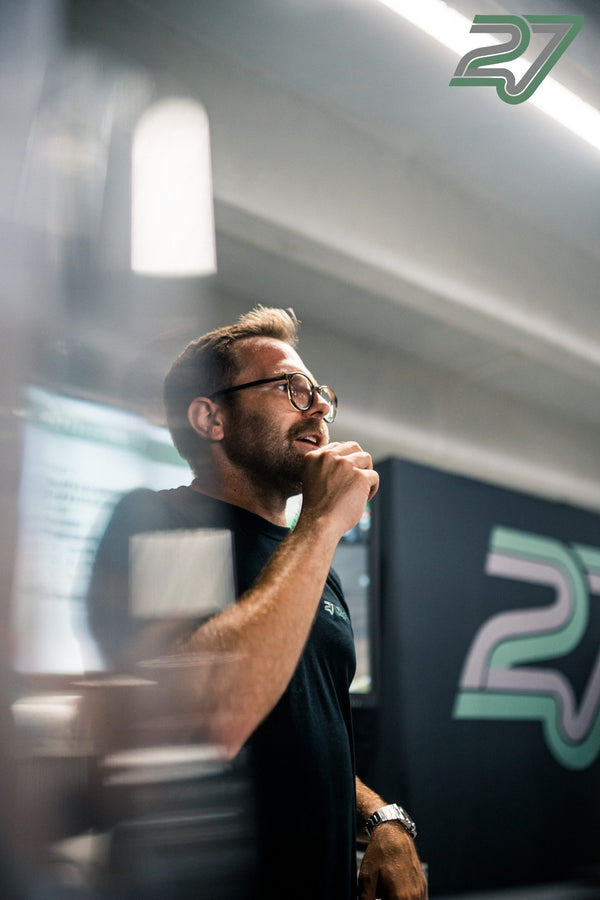 Bärtiger Mann mit Brille spricht gestikulierend vor einem beleuchteten 27-Logo