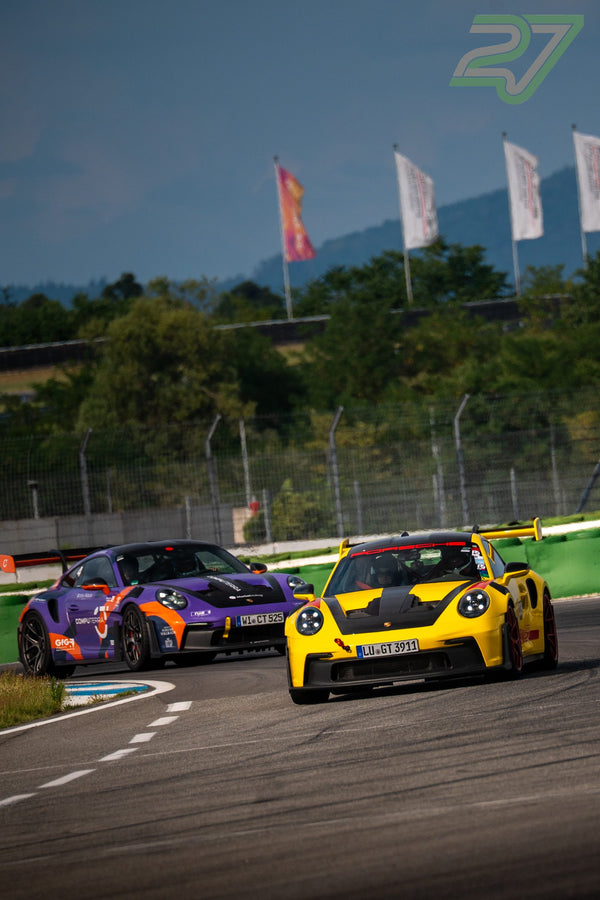 Gelber Porsche GT3 führt einen lila-orangen Porsche GT3 durch die Kurve vor dunklen Wolken