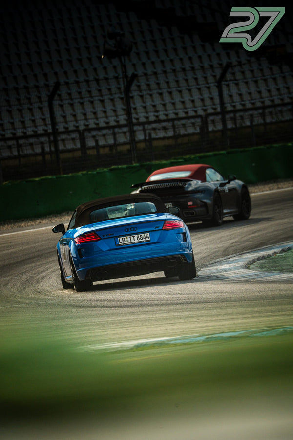 Blauer Audi TT Roadster mit geschlossenem Verdeck fährt vor einem dunklen Porsche in die Kurve