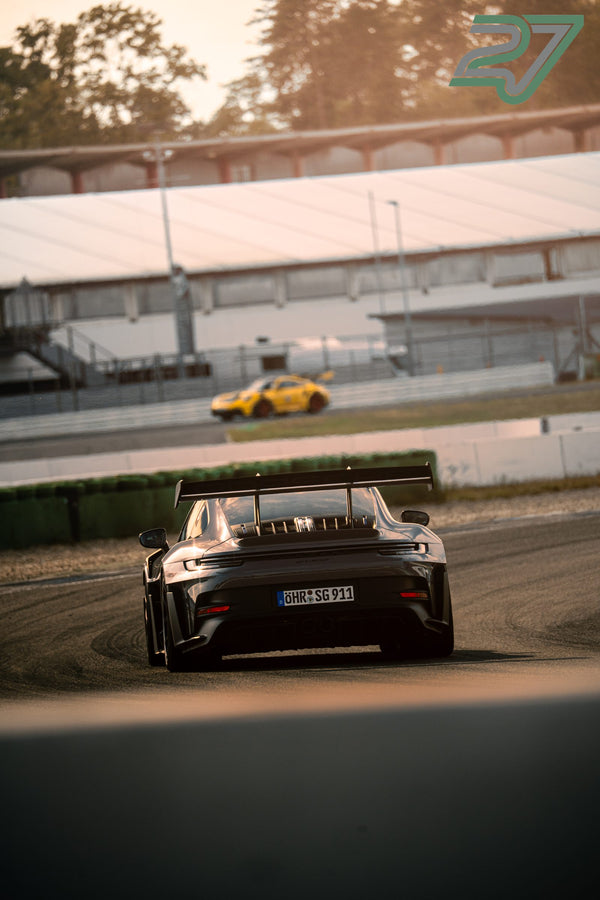 Dunkler Porsche mit großem Heckflügel fährt im goldenen Licht, im Hintergrund ein verschwommener gelber Porsche