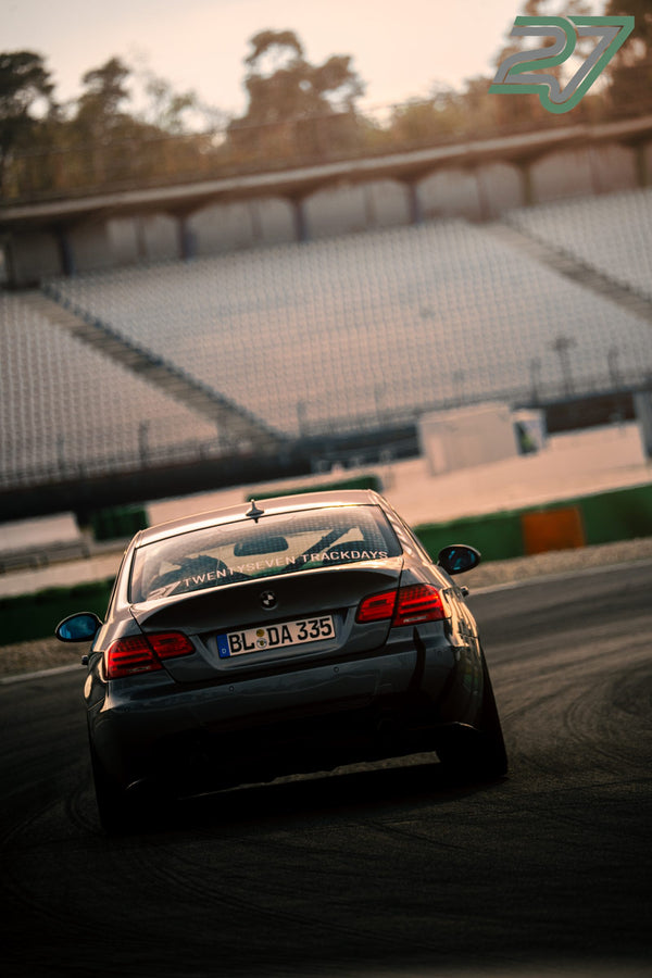 Dunkler BMW 3er-Coupé mit Trackdays-Schriftzug im Heckfenster fährt im warmen Abendlicht davon