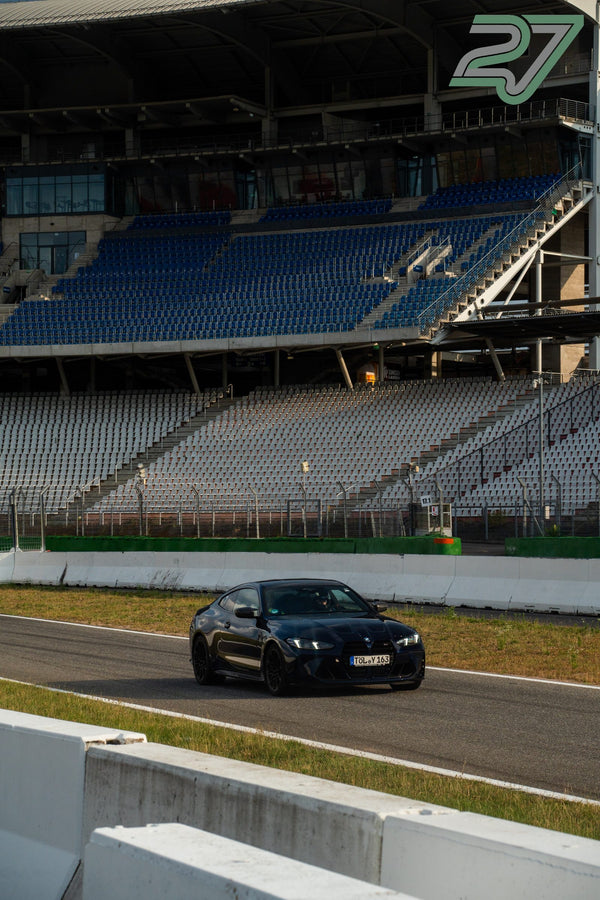Dunkler BMW fährt im Abendlicht frontal auf die Kamera zu, dahinter die leere Tribüne des Hockenheimrings