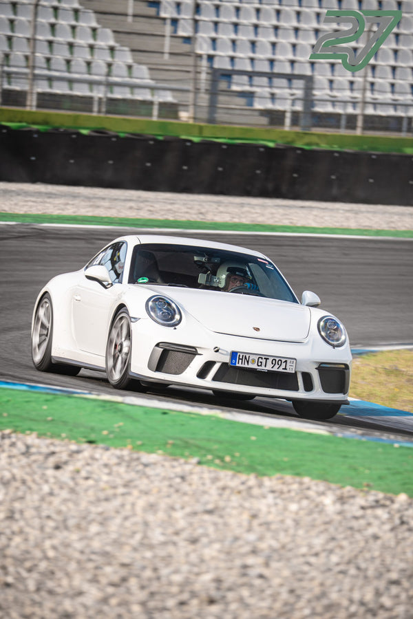Weißer Porsche 911 GT3 mit zwei Insassen fährt in einer Kurve über den grünen Randstein