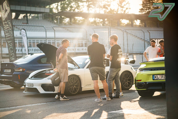 Drei Männer unterhalten sich vor einem weißen Porsche mit offener Fronthaube im Gegenlicht