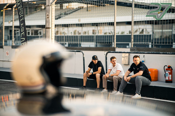 Drei Männer sitzen locker im Gespräch auf der Boxenmauer, im Vordergrund ein unscharfer Helm