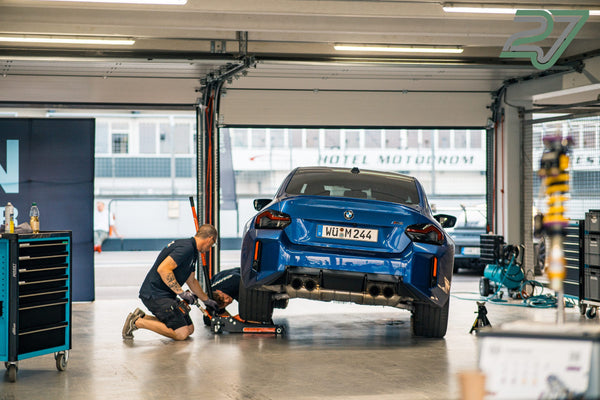Zwei Mechaniker arbeiten an einem aufgebockten blauen BMW M2 vor dem Schriftzug Hotel Motodrom