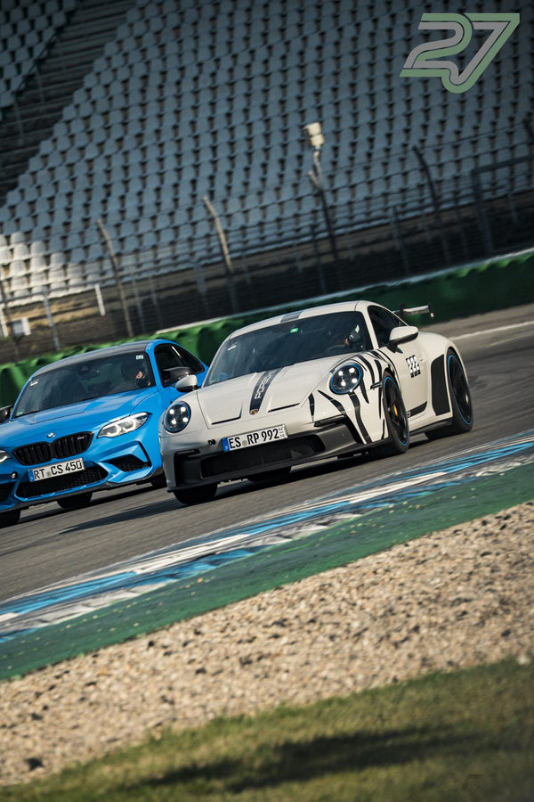 Weißer Porsche 911 GT3 mit Rennstreifen fährt neben blauem BMW M2 durch die Kurve