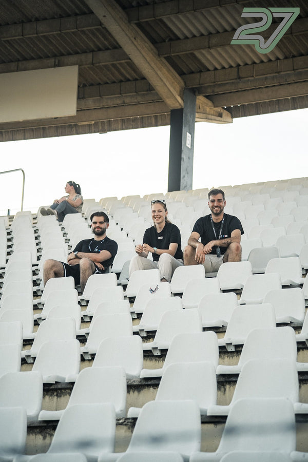 Drei Personen sitzen lächelnd nebeneinander in der leeren Tribüne und blicken in die Kamera
