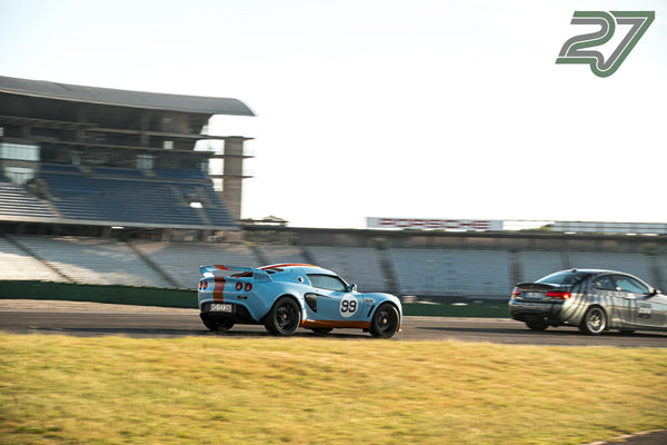 Blau-oranger Lotus Exige biegt vor einem dunklen Wagen mit Tarnmuster in die Kurve