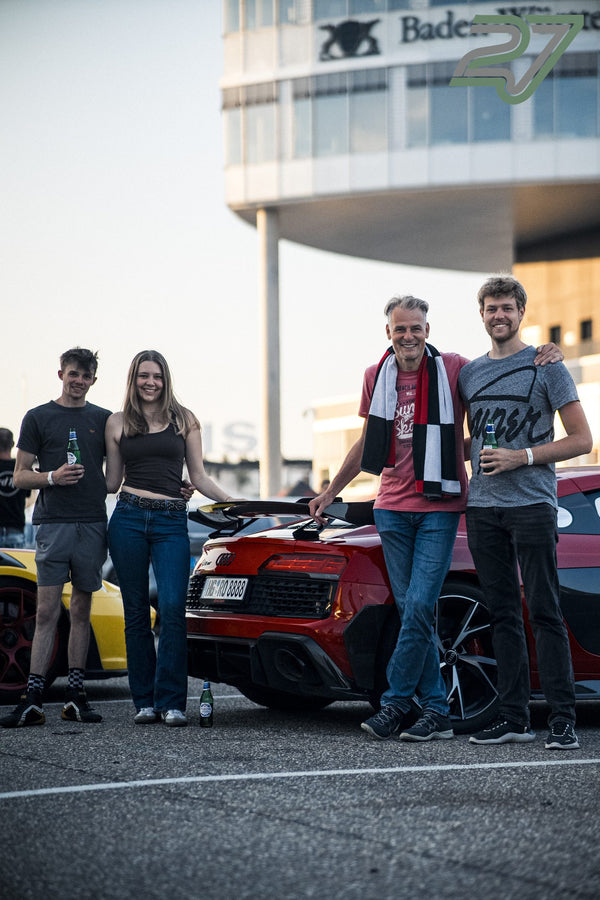 Vier lächelnde Teilnehmer posieren mit Getränken vor einem roten Audi R8 und einem Turm