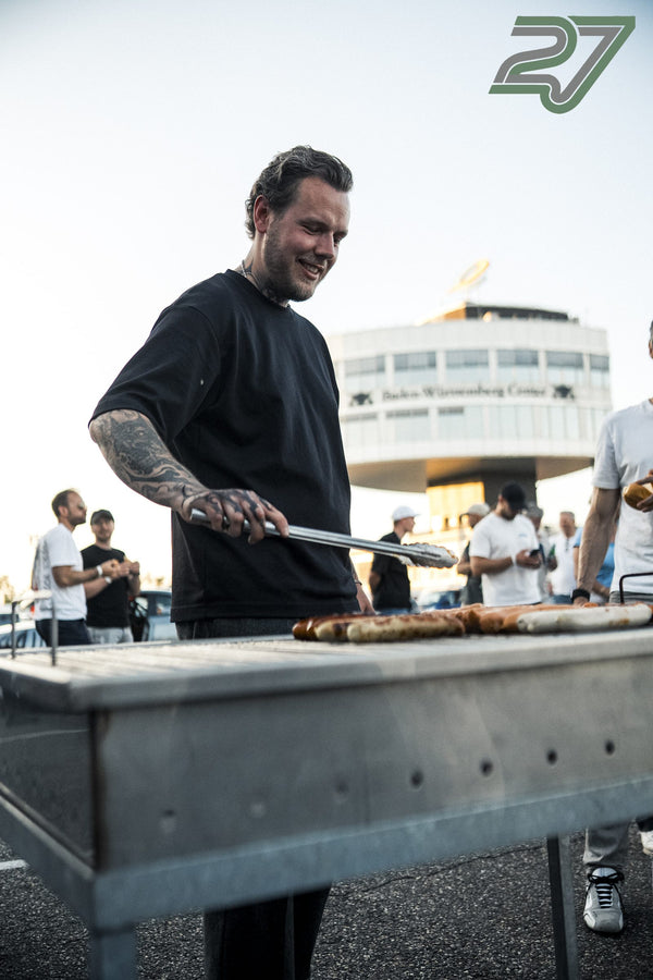 Tätowierter Mann wendet lächelnd Würstchen auf dem Grill, im Hintergrund ein Turm