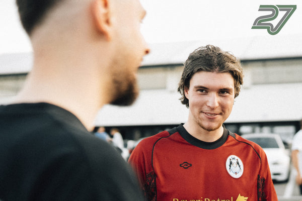 Zwei Männer plaudern entspannt, einer trägt ein rotes Sport-Trikot mit Wappen
