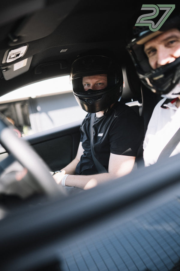 Zwei Fahrer mit Helm sitzen angeschnallt nebeneinander im Auto, einer lächelt breit