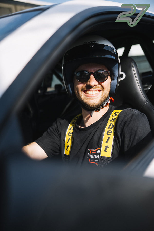 Fahrer mit offenem Helm und Sonnenbrille lacht breit, der gelbe Renngurt ist sichtbar
