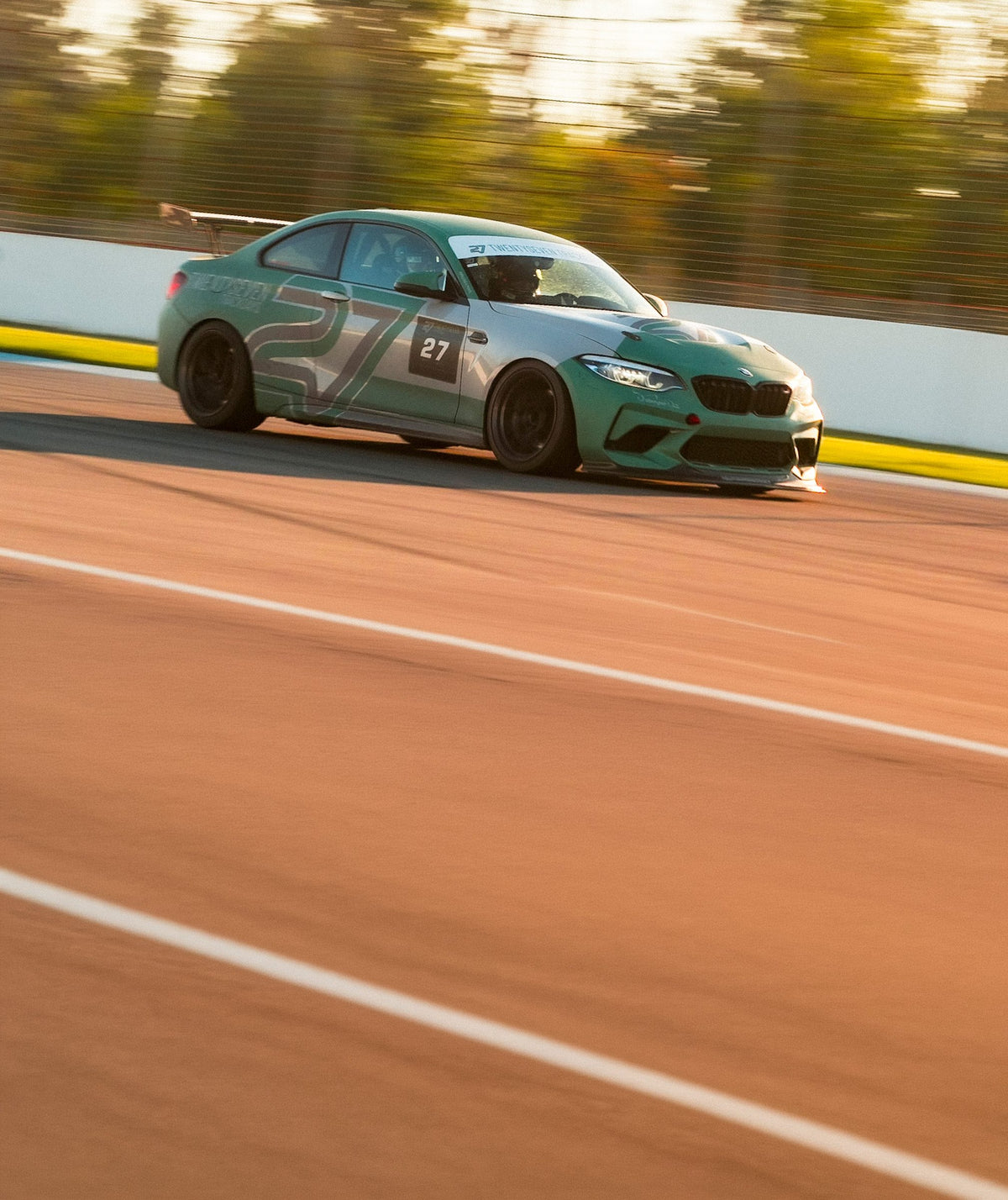 24/7 Performance BMW M2 Competition in the green and grey TWENTYSEVEN livery with race number 27, front view backlit by the evening sun on the circuit