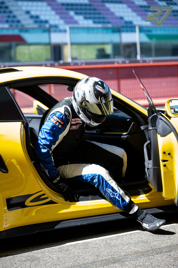 Im blau-weißen Overall steigt ein behelmter Fahrer aus dem gelben Porsche, Tribüne im Rücken