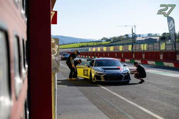 Gelb-silberner Audi R8 mit Startnummer 27 in der Boxengasse, drei Helfer am Auto, Hügel dahinter