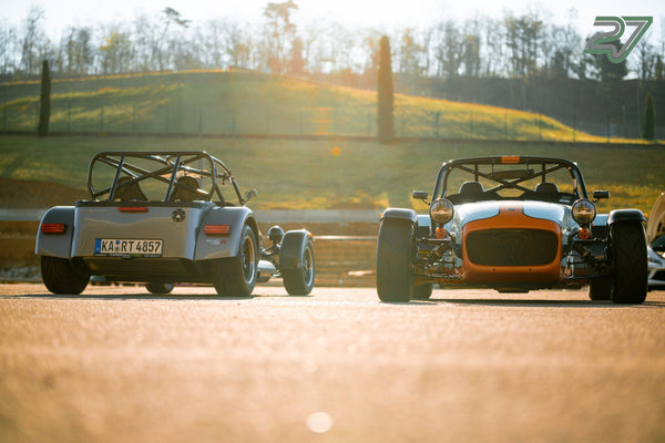 Zwei Caterham Seven im Gegenlicht, einer von hinten, einer von vorn, dahinter ein begrünter Hang
