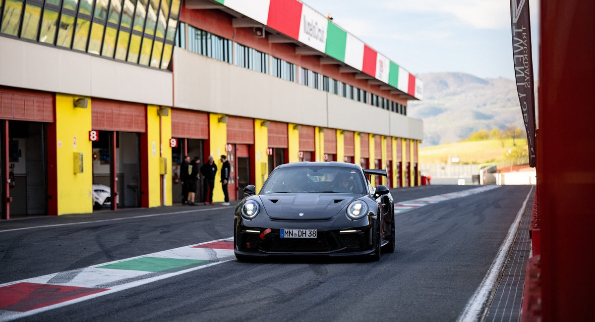 Schwarzer Porsche 911 GT3 RS rollt die Boxengasse entlang, Trikolore am Gebäude, Berge im Rücken