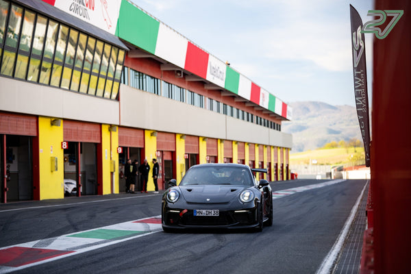 Schwarzer Porsche 911 GT3 RS rollt die Boxengasse entlang, Trikolore am Gebäude, Berge im Rücken