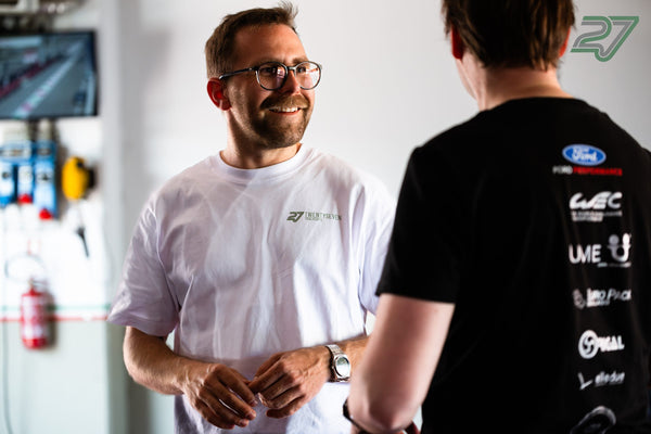 Mann mit Brille im weißen TWENTYSEVEN-Shirt lacht im Gespräch mit einer Person in schwarzem Shirt
