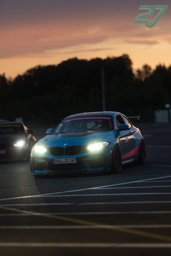 Türkisblauer BMW M2 mit pinkem Streifen und leuchtenden Scheinwerfern rollt bei Sonnenuntergang ins Fahrerlager