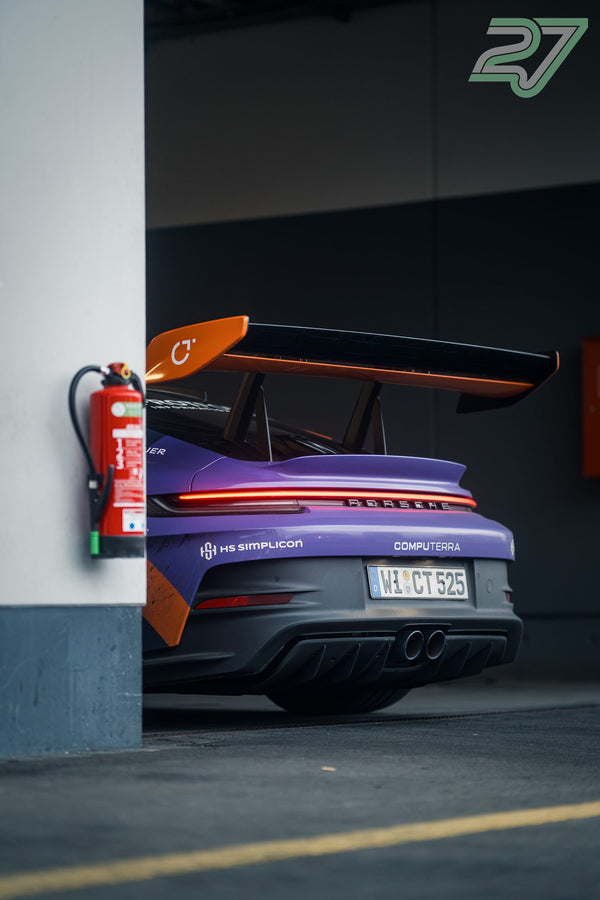 Violetter Porsche mit großem orangefarbenem Heckflügel steht in der Box neben einem Feuerlöscher