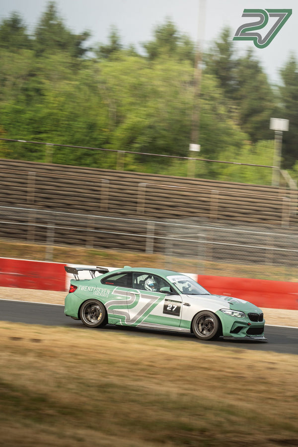 Mint-grüner BMW M2 mit Twentyseven-Beschriftung rast vor grüner Waldkulisse über die Strecke