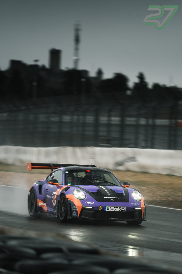 Violett-oranger Porsche 911 GT3 driftet mit Wasserfontäne vor der Nürburg durch die Kurve