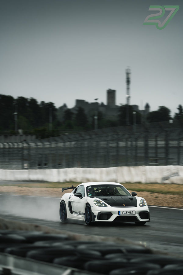 Weißer Porsche Cayman GT4 RS zieht mit feiner Gischt vor der Burgkulisse durch die nasse Kurve