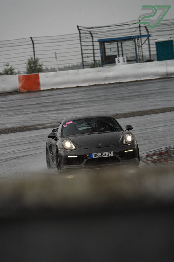 Dunkler Porsche Cayman fährt mit eingeschalteten Scheinwerfern über die spiegelnd nasse Fahrbahn