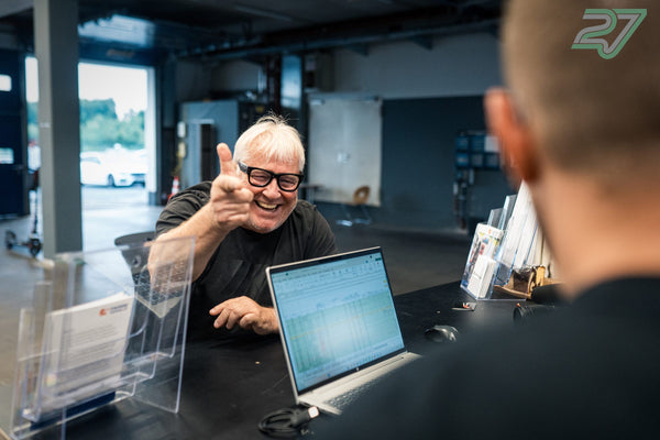 Mann mit weißem Haar und Brille lacht und deutet mit dem Finger zur Kamera am Empfangstisch