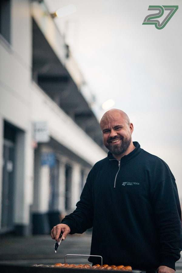 Mann im schwarzen Trackday-Pullover grillt lächelnd Würstchen im Abendlicht
