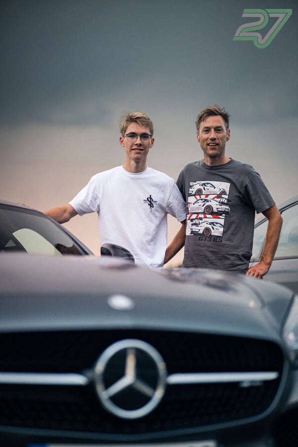 Zwei Männer stehen lächelnd neben grauem Mercedes-Sportwagen bei bewölktem Himmel