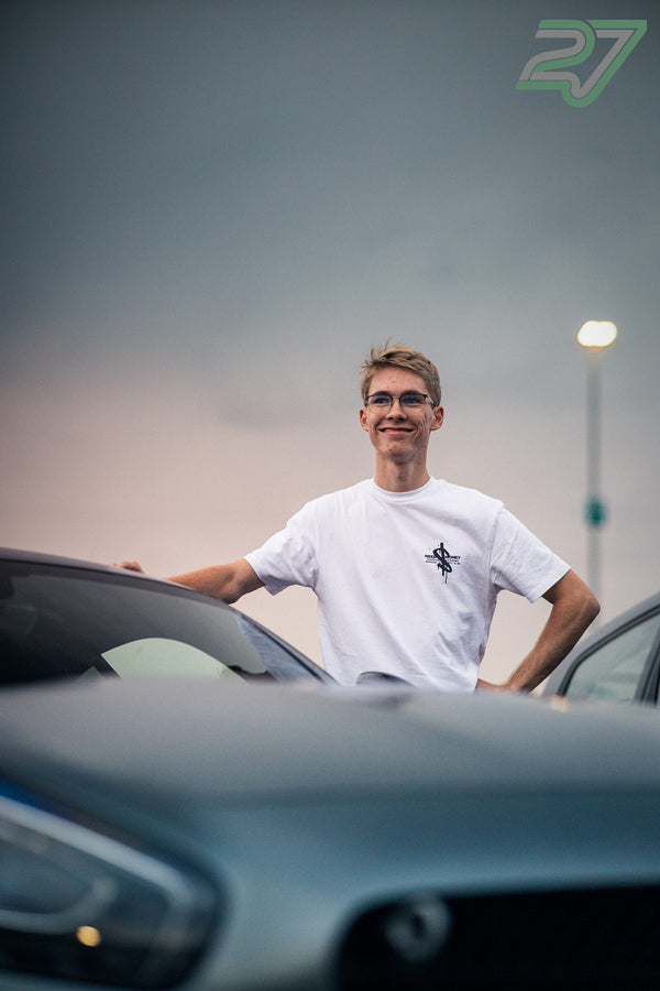 Mann mit Brille steht lächelnd zwischen parkenden Autos in der Abenddämmerung auf dem Parkplatz