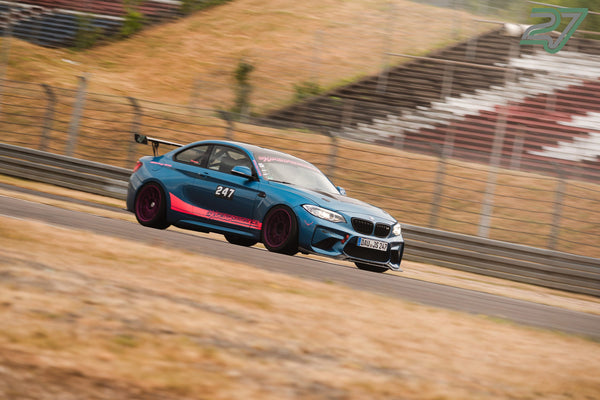 Türkis-pinkfarbener BMW M2 rast mit Überrollkäfig durch eine Kurve auf der Rennstrecke