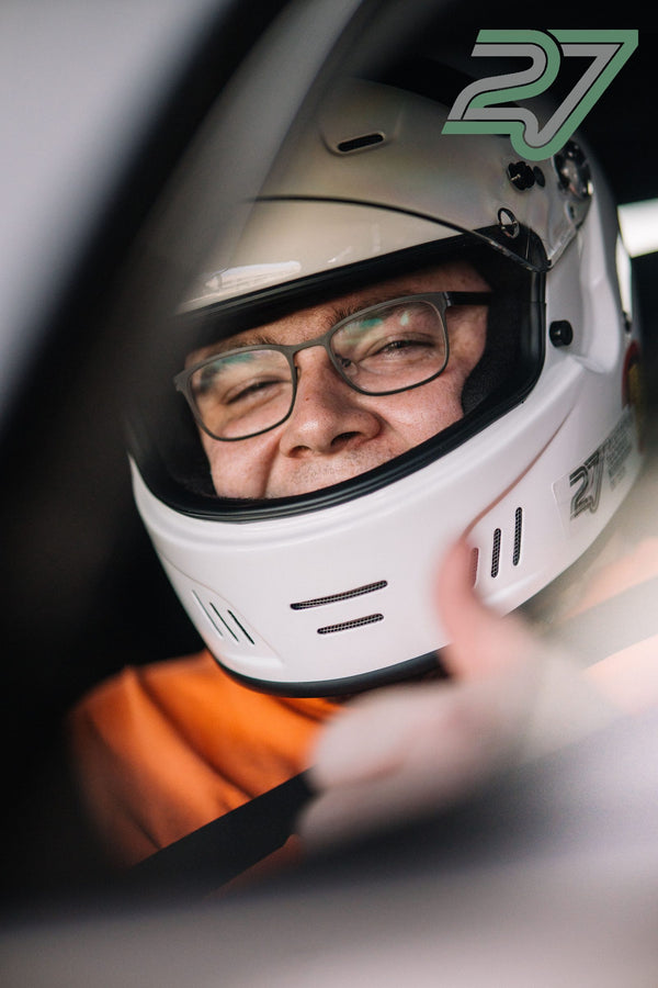 Fahrer mit weißem Helm sitzt angeschnallt im Cockpit und hält lächelnd den Kinnriemen fest