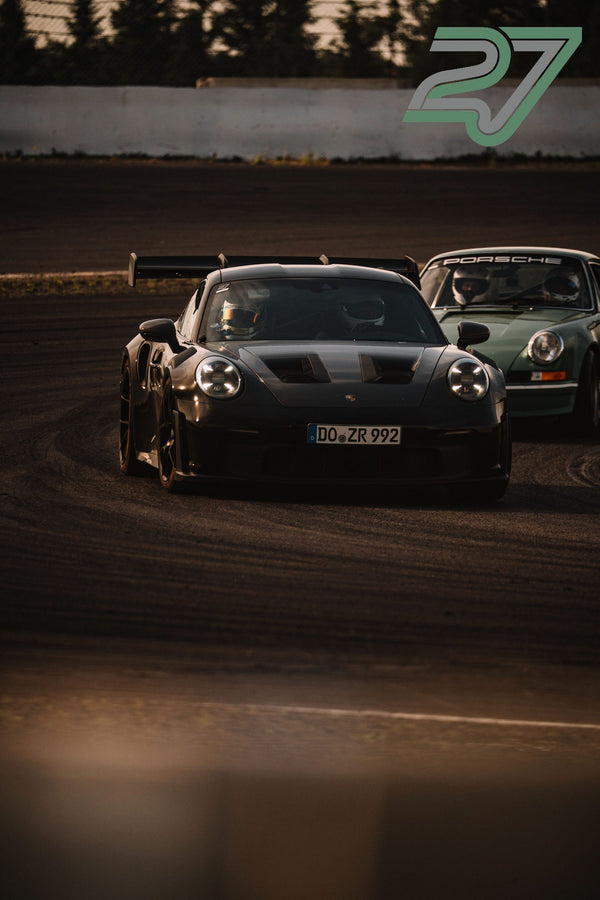 Schwarzer und mintgrüner Porsche 911 fahren hintereinander im warmen Abendlicht durch eine Kurve