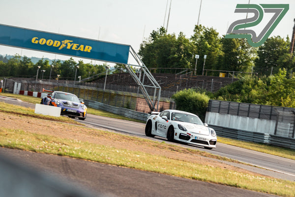 Weißer Porsche Cayman fährt scharf gezeichnet durch eine Kurve am Nürburgring, dahinter ein verschwommener Porsche 911