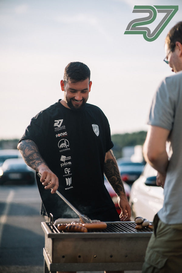 Tätowierter Mann lacht beim Grillen von Bratwürsten, im Hintergrund eine zweite Person