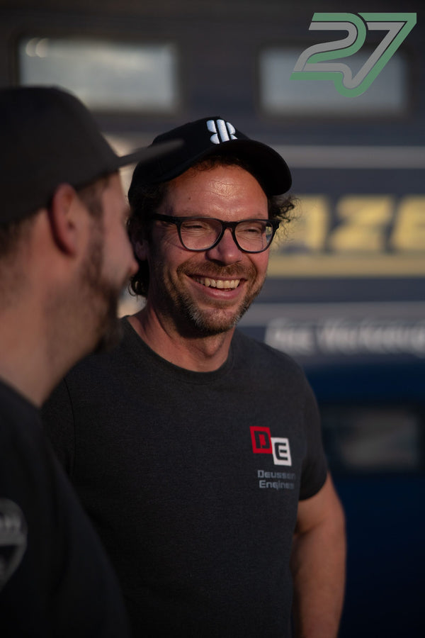 Zwei Männer lachen gemeinsam vor einem Transporter im warmen Abendlicht des Fahrerlagers