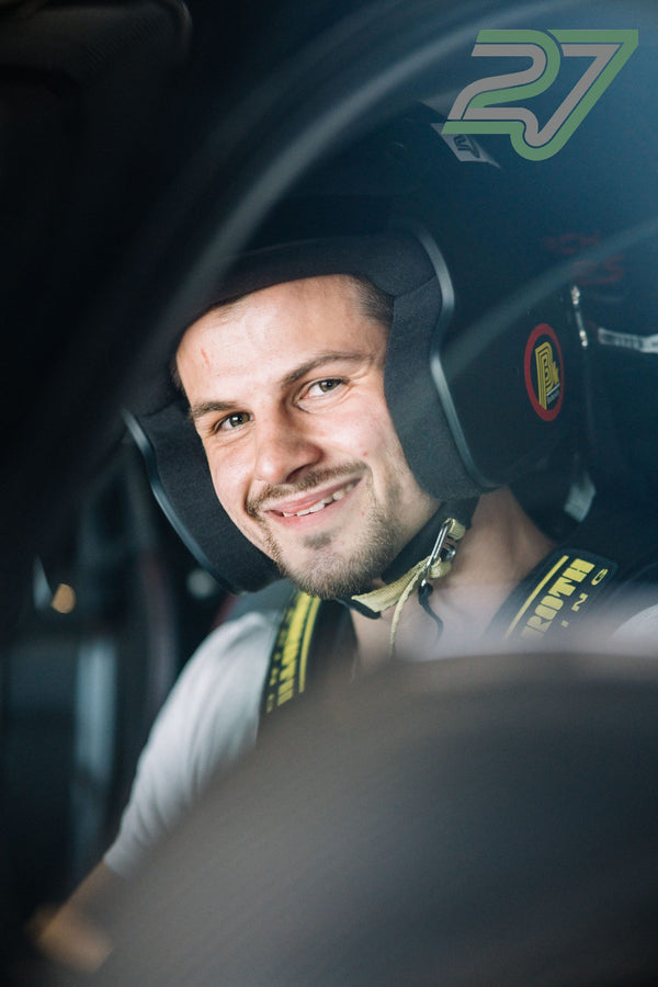 Junger Mann mit dunklem Helm und offenem Visier lächelt breit angeschnallt im Auto