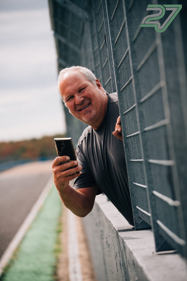 Älterer Zuschauer lehnt lachend durch die Streckenbegrenzung und fotografiert mit dem Smartphone