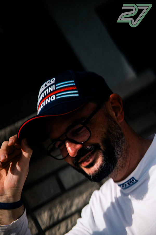 Mann mit Sparco-Martini-Racing-Cap lüftet lachend den Schirm im warmen Gegenlicht