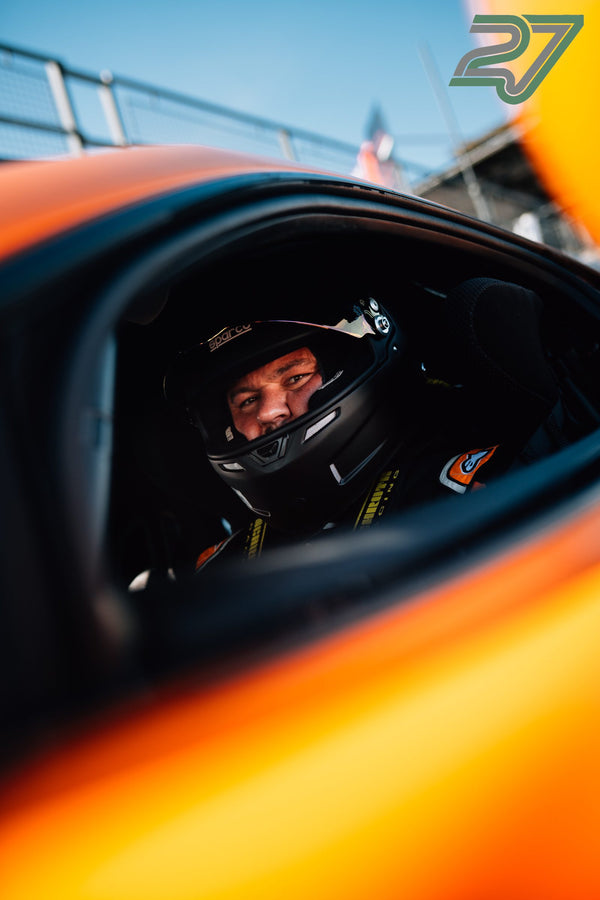 Fahrer mit Helm sitzt im orangefarbenen Cockpit, gespiegelt im Außenspiegel des Wagens