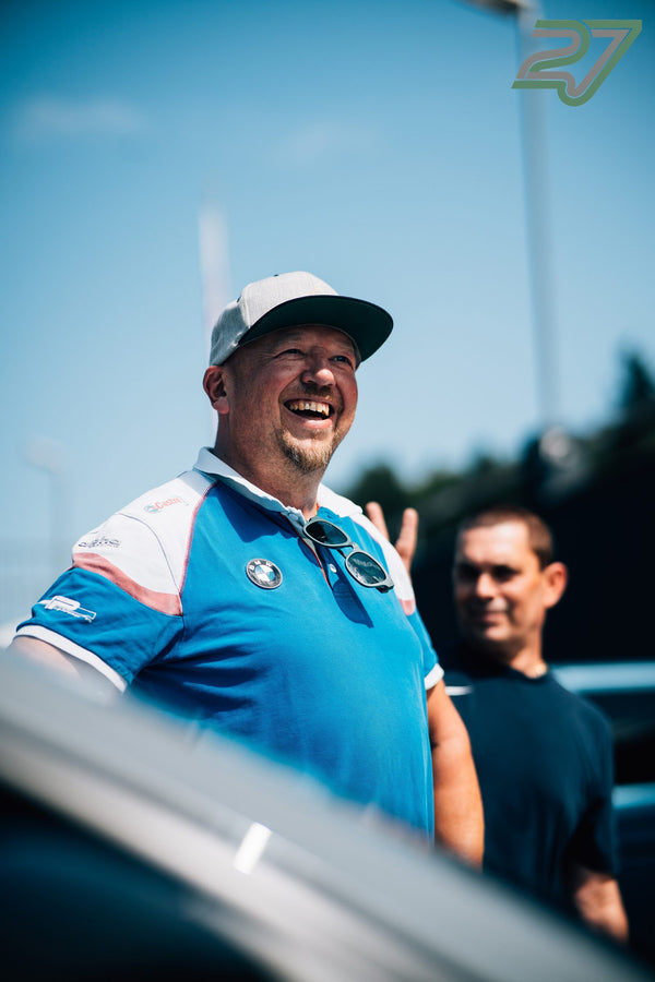 Lachender Mann mit grauer Cap und hellblauem BMW-Poloshirt zeigt breit grinsend das Peace-Zeichen