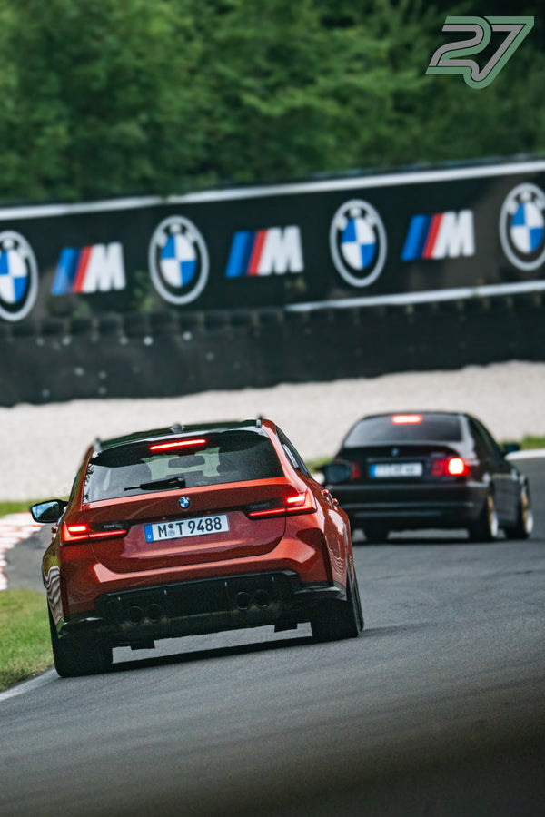 Oranger BMW M3 führt vor einem schwarzen BMW an der BMW-M-Bannerwand im Wald vorbei