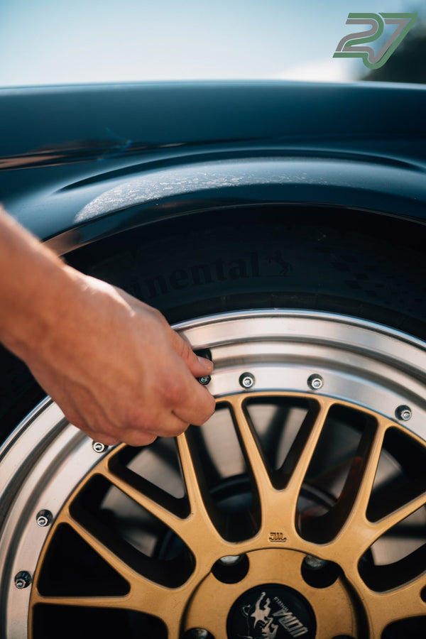 Nahaufnahme einer Hand, die eine goldene Leichtmetallfelge mit BMW-Nabendeckel am petrolfarbenen Auto montiert