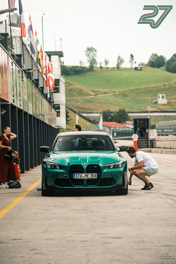Ein Mann kniet am Vorderrad des türkisen BMW M3, eine Frau im weinroten Kleid wartet mit Helm
