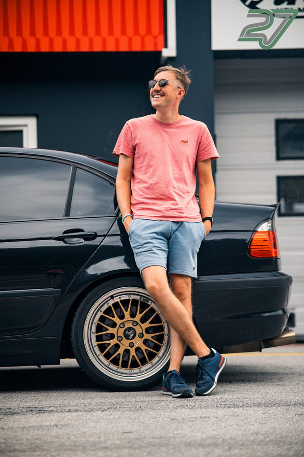 Ein Mann im korallenfarbenen T-Shirt lehnt lachend an einem schwarzen BMW mit goldenen Felgen
